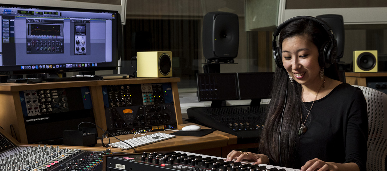 A woman wearing a black shirt practices piano while wearing headphones in a music engineering studio.